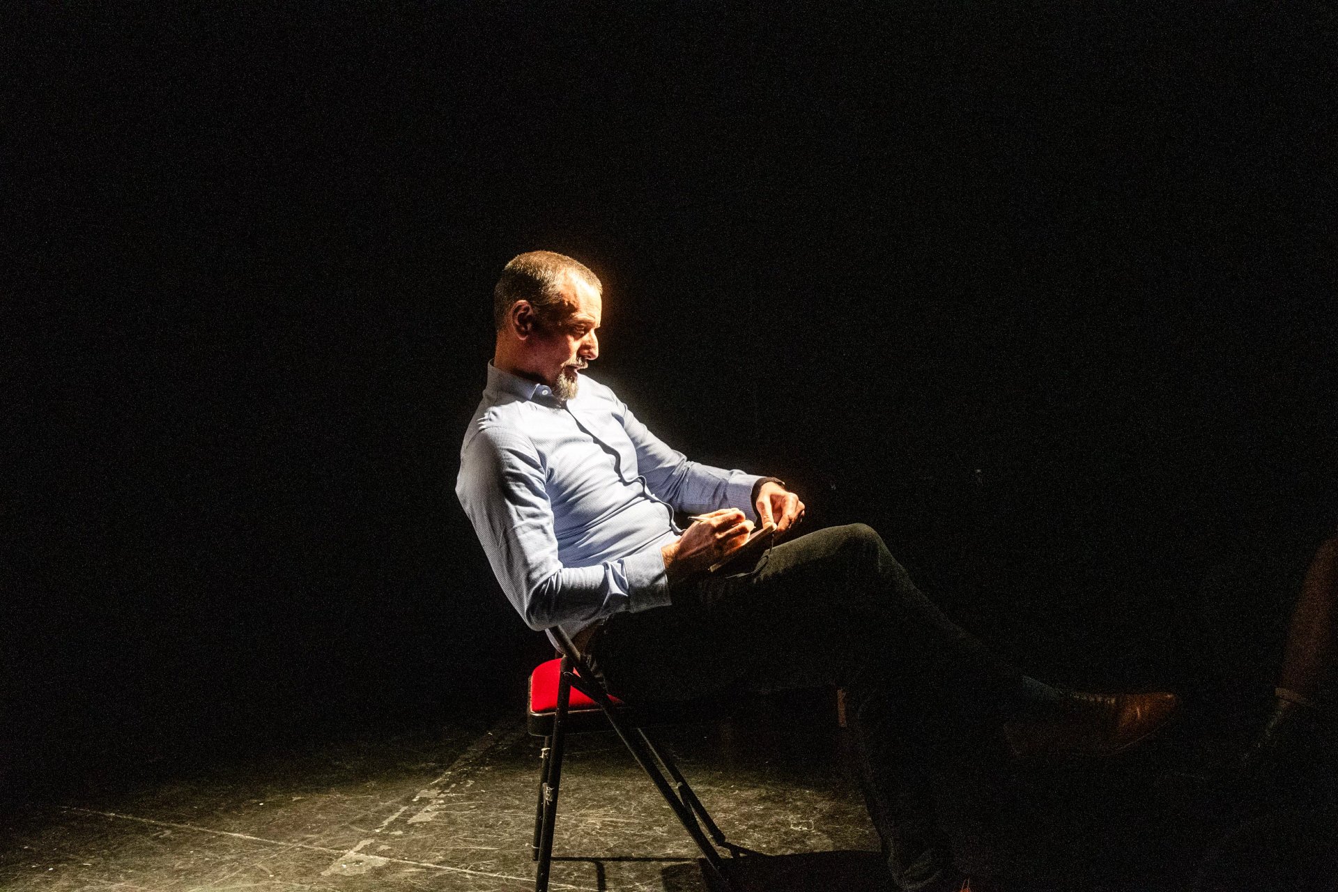 An actor sits onstage writing in a book. He is lit in a spotlight and is wearing a shirt and trousers. He looks contemplative.