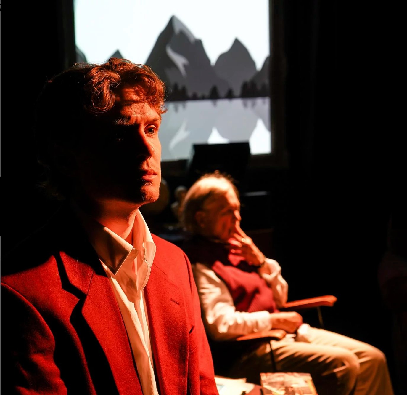 A woman sits in the background and a man sits in the foreground. They are lit in an orange light that only lights the right side of them leaving the rest in shadow. A black and white image is projected behind them of mountains.