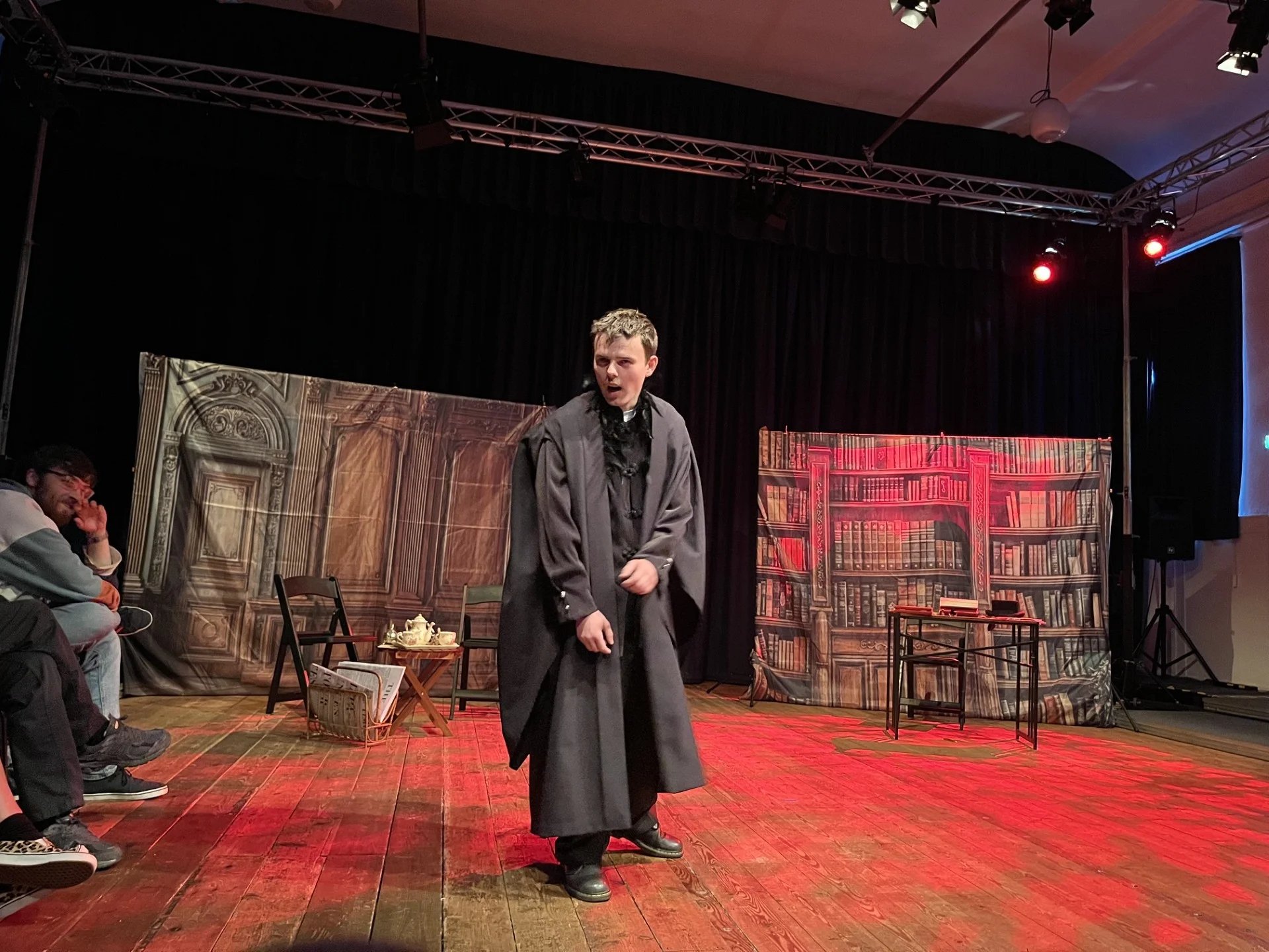 An actor in a black robe stands bent over on stage. There is a red light on the floor behind him in a blood splatter like pattern.