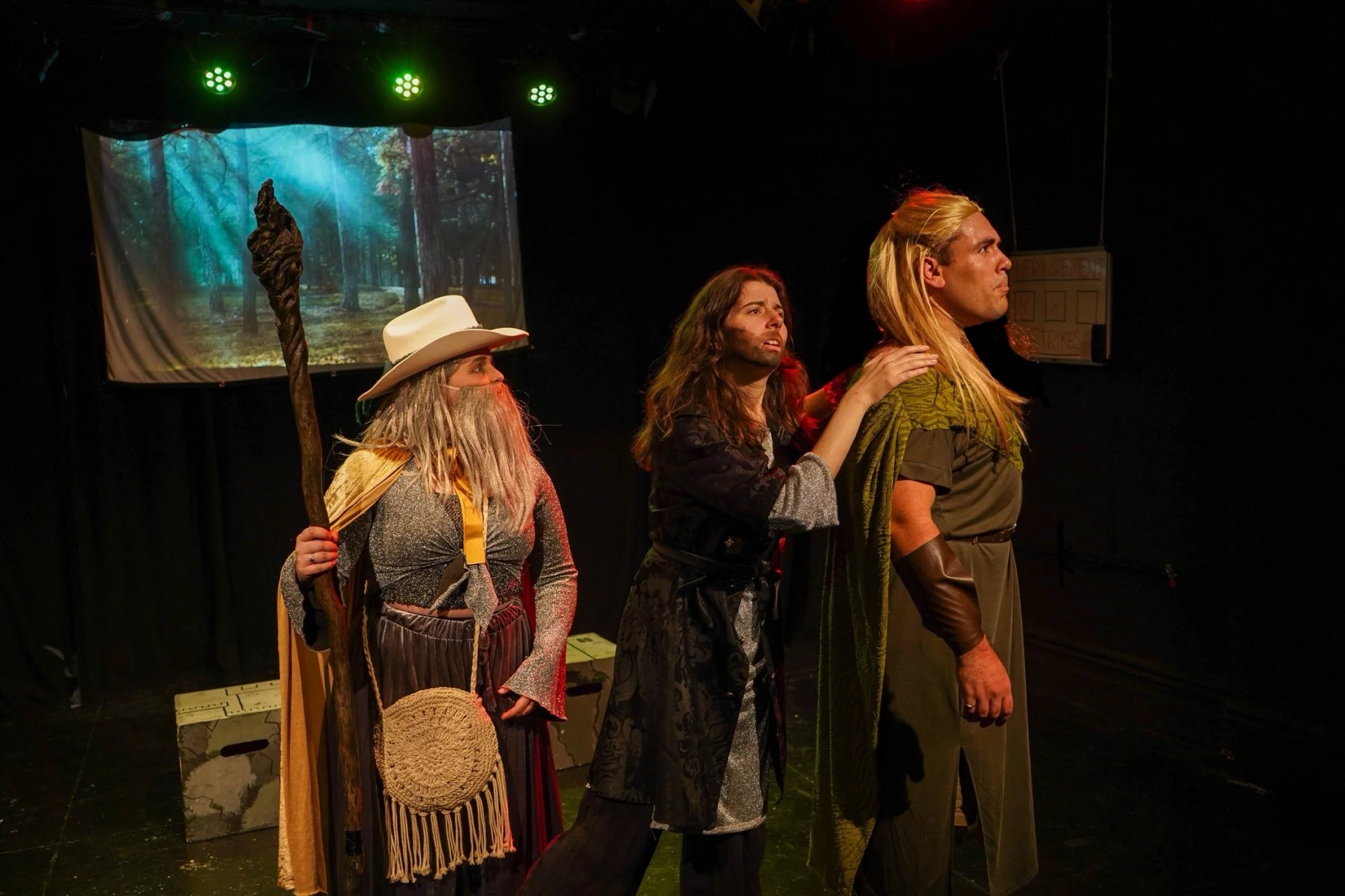 Three actors stand on stage looking to the right. A red light glows on them. One is wearing a cowboy hat and gold cloak holding a tall wooden staff. Another is all in black. The third is wearing a green cloak and dress.