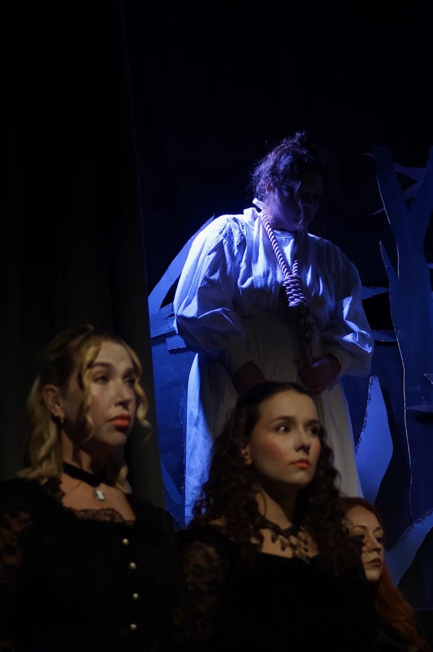The image focuses on a woman in a white dress with a noose around her neck, she is lit in white light with strong shadows. In the foreground is the out of focus faces of concerned actors lit in yellow.