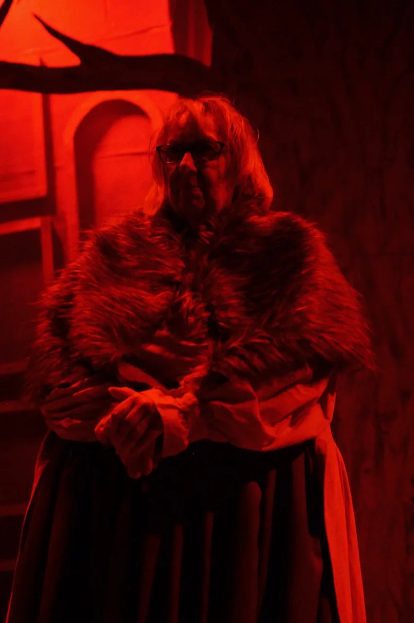 A woman in a fur shawl stands in front of a tree. The whole image is lit by red light.