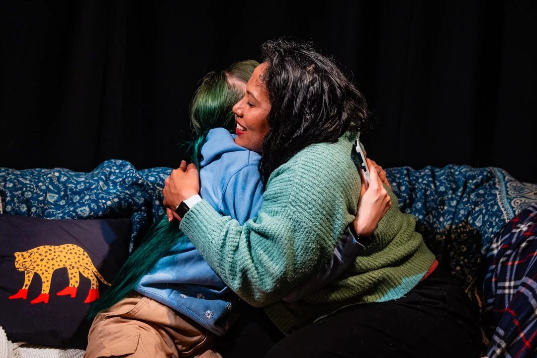 Two actors hug each other while sat on a sofa. They are wearing a hoodie and a cardigan, and look happy.