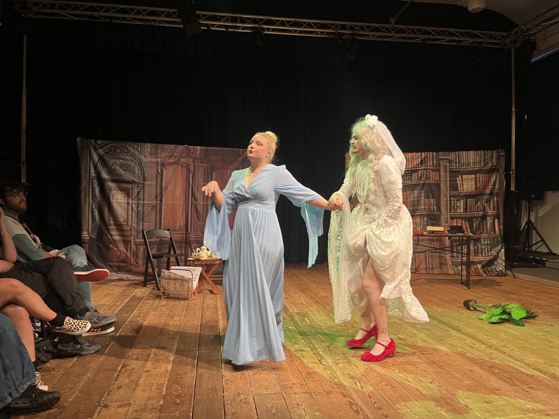 An actor in a blue dress leads another in a wedding dress and red shoes across stage. The lighting is warm and there is some green with it.