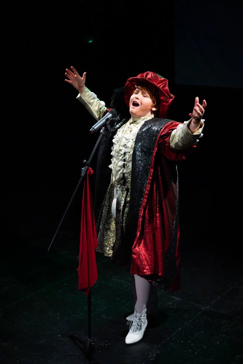 Hannah Clift is dressed as Henry the eighth. They are singing into a microphone with arms wide.