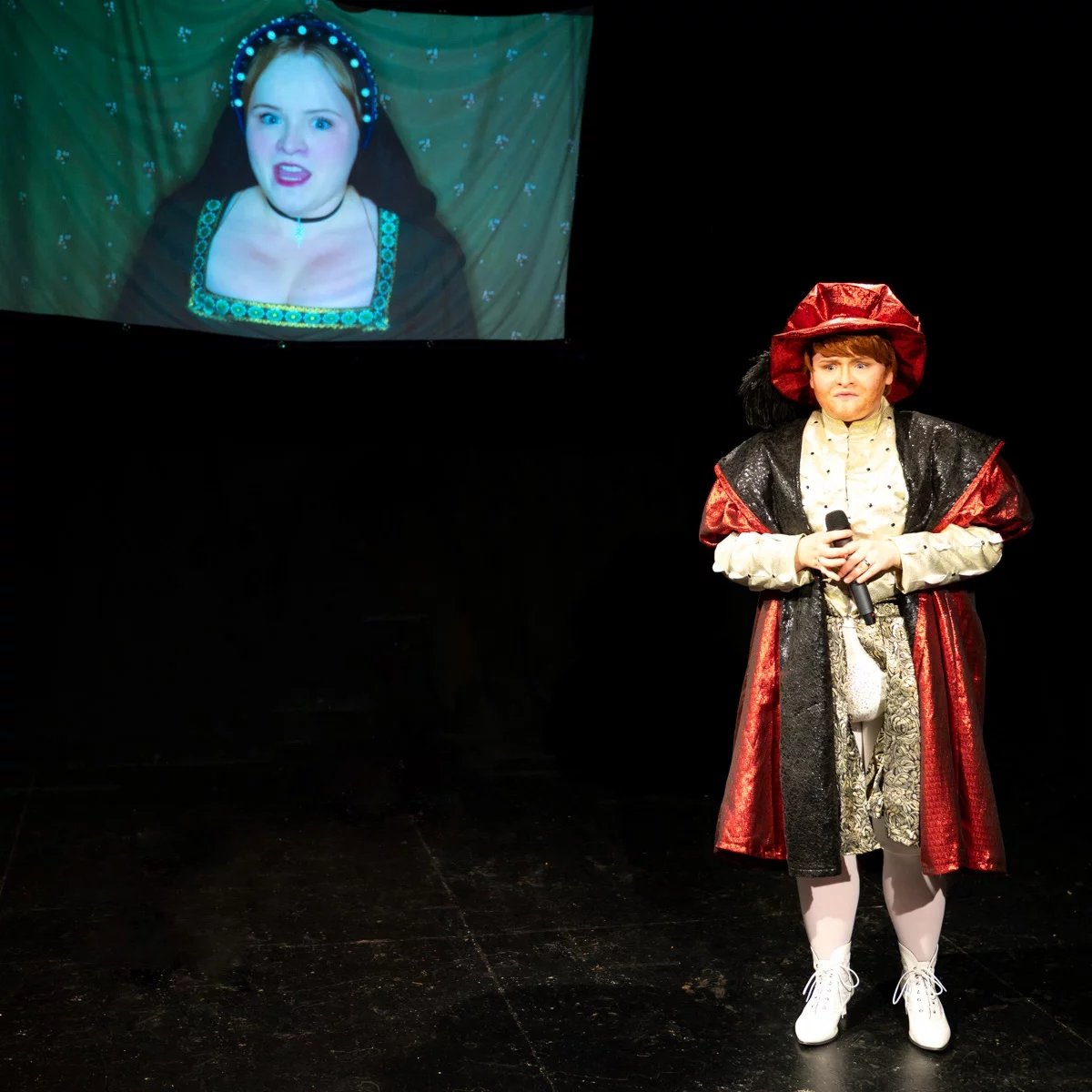 Henry the eighth stands in the foreground holding a microphone looking unhappy. In the background a projected image shows an actor as Anne Boleyn singing.