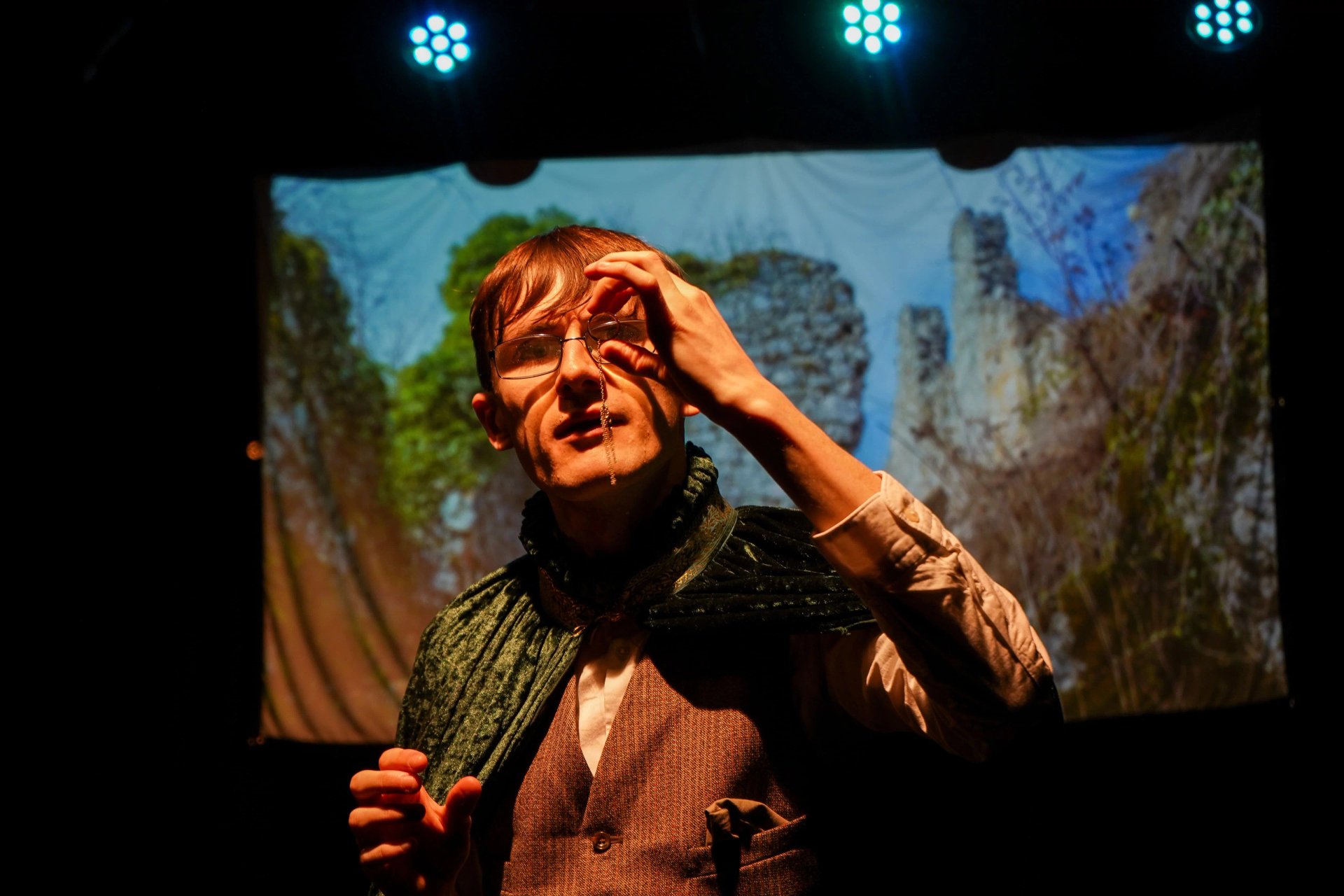 A man stands in orange light holding a ring in front of his eye. He is wearing a green cloak, brown waist coat and white shirt. Behind him is a projected image of ruins.