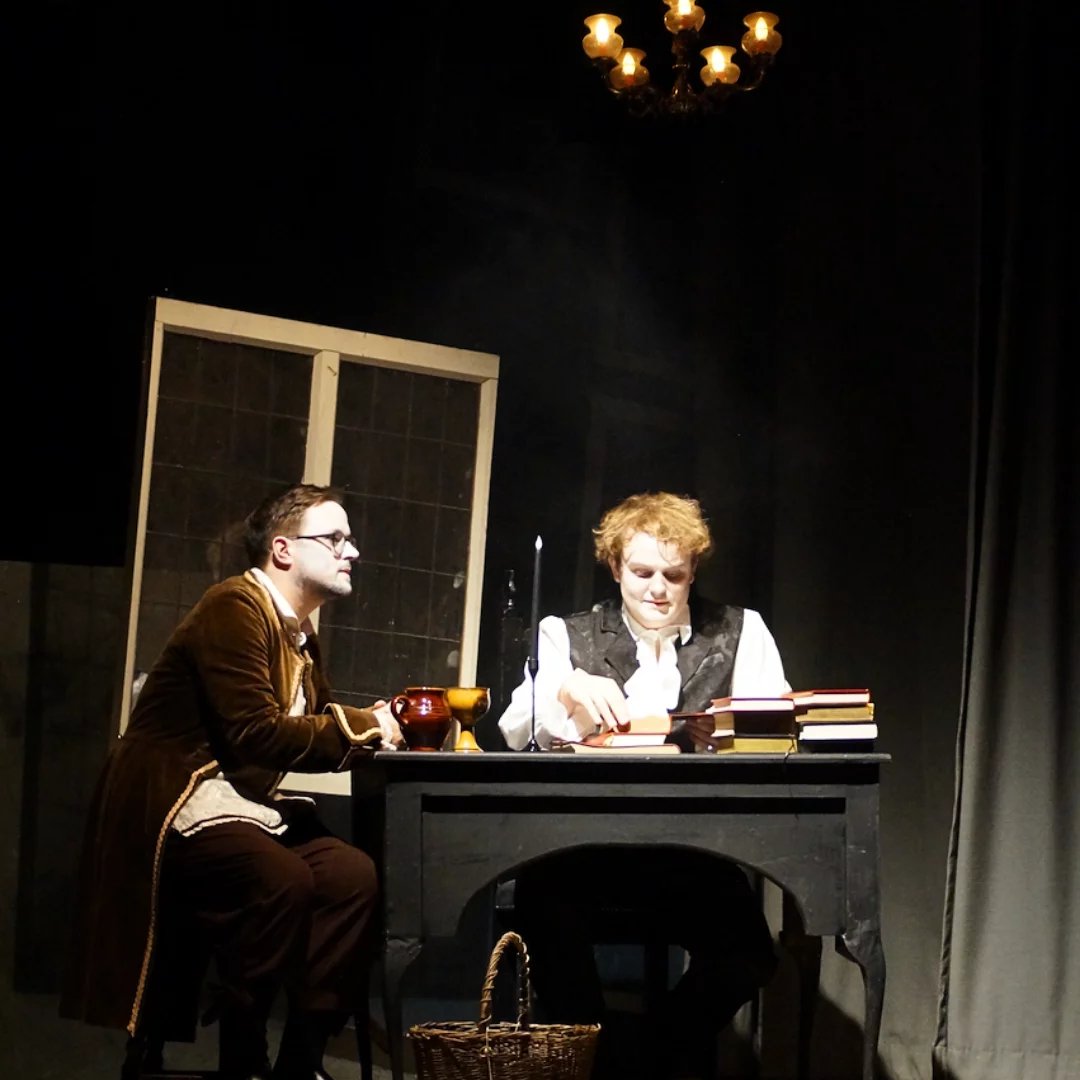 Two men sit at a desk writing. There is a window behind them and a chandelier lit above them. They have white painted faces and are in 1800's clothing.