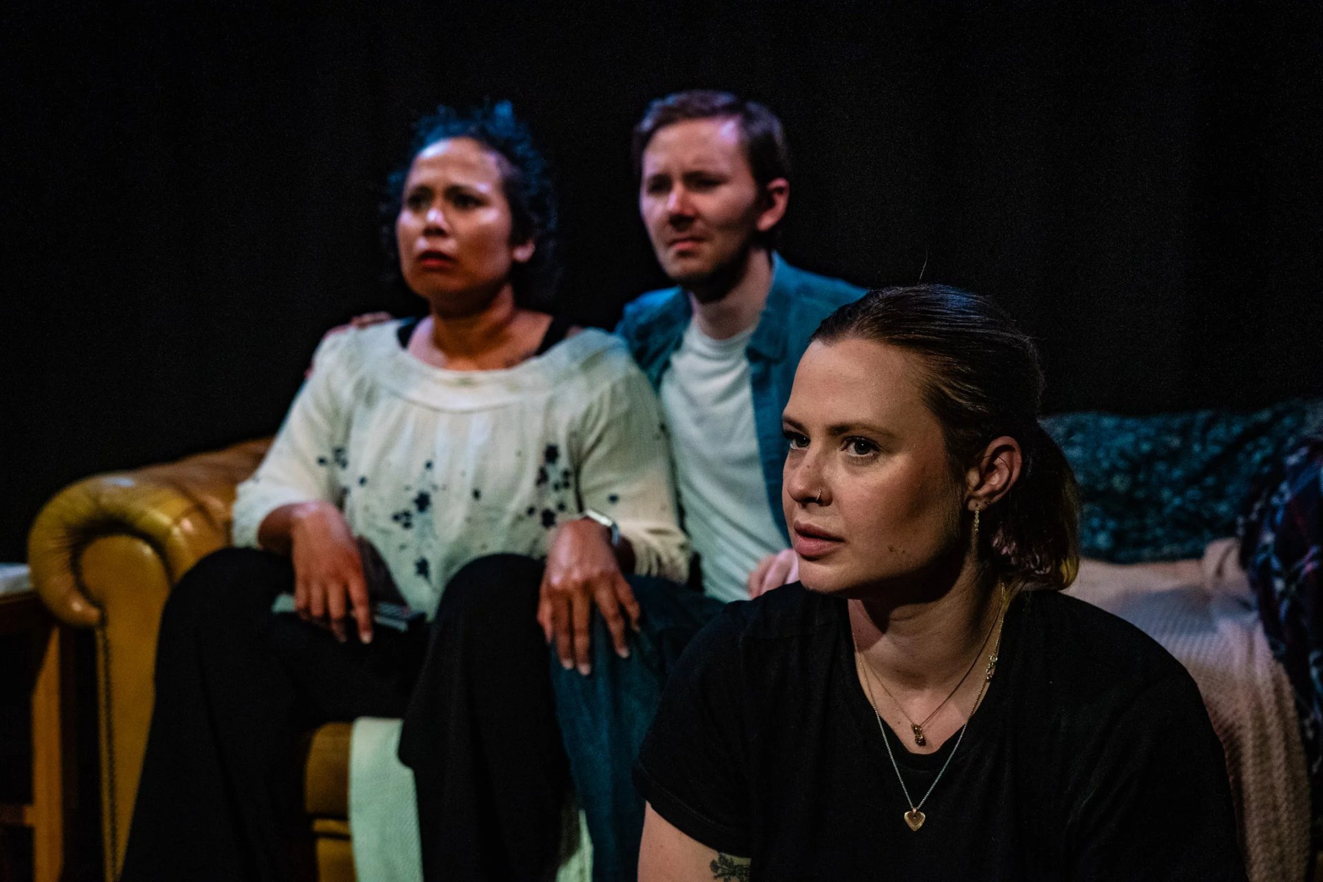 Two actors sit on a sofa and one on the floor in front of them. They are in low lighting and look concerned and upset.