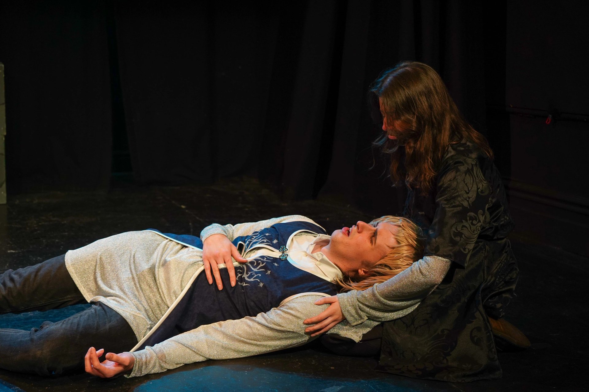 A man lies in another's lap holds his chest and looking in pain. He is wearing chainmail and a blue surcoat. The other man is all in black, their hair obscuring their face.