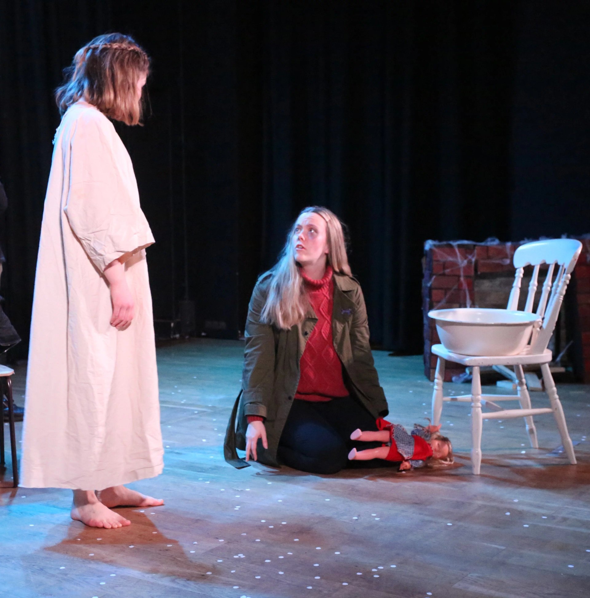A woman sits on the floor with a doll beside her. Another woman in a white shroud stands over her. The lighting is white and blue.