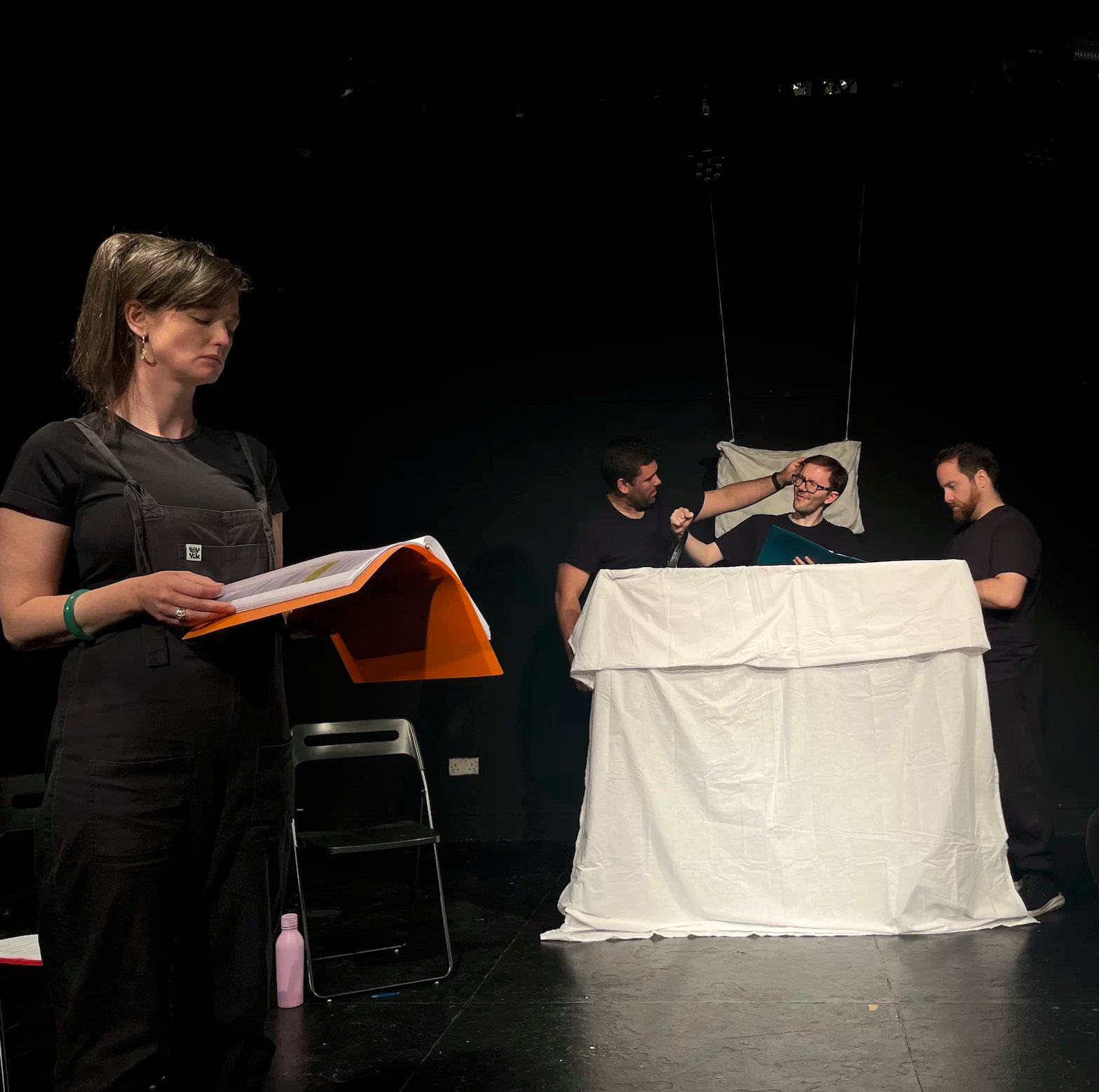 An actor stands in a black theatre holding a script. Three actors are in the background of the image behind a white sheet that is being used as a bed.