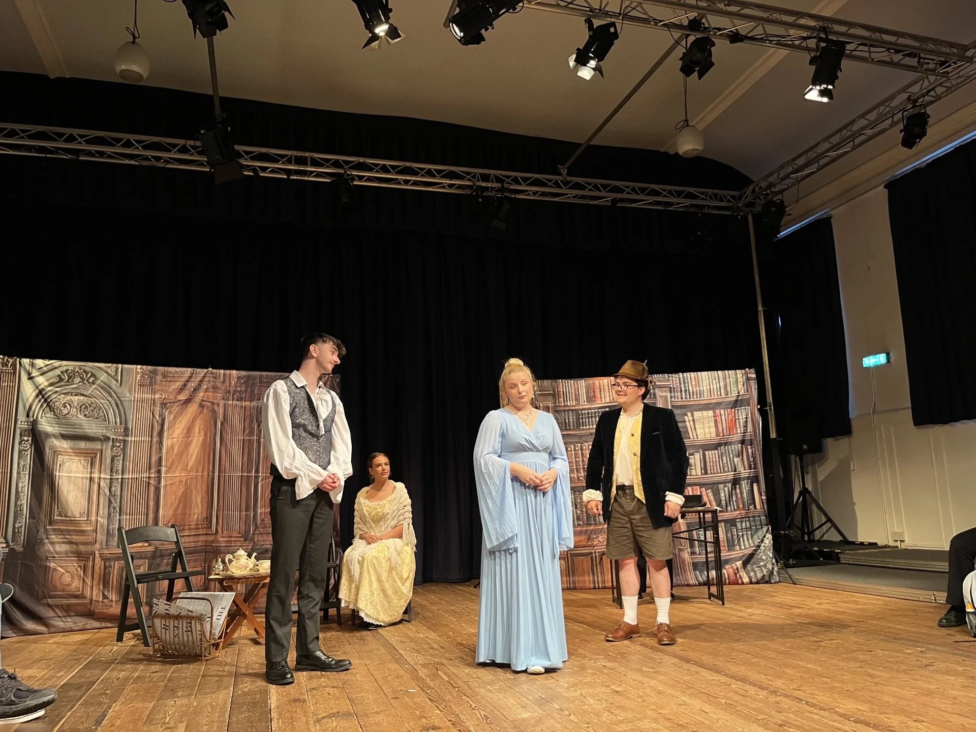 Three actors stand onstage, another is sitting. They are in 19th century clothing and lit by a warm lighting.