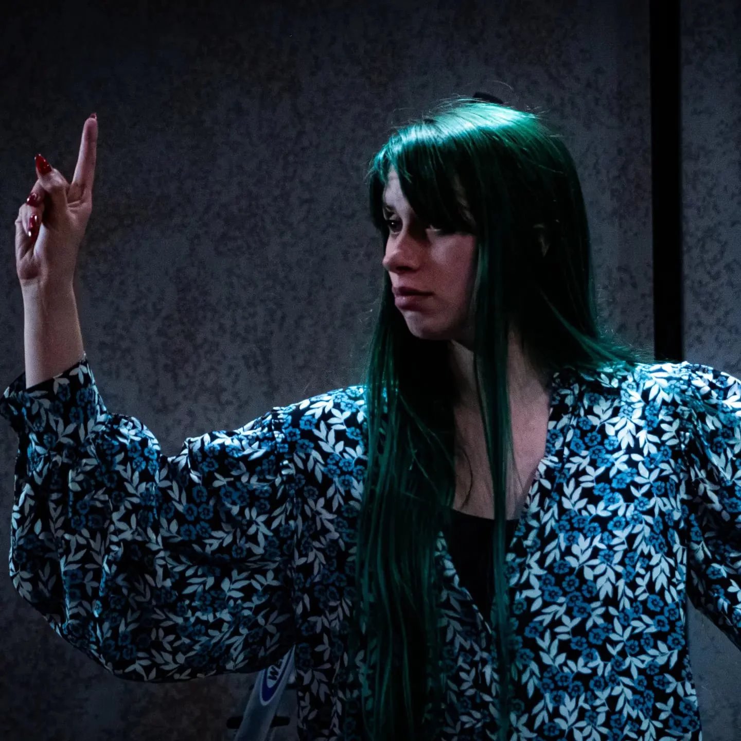 Billie-Jo as the fool in Queen Lear. She has a floral shirt on and is holding a finger in the air.