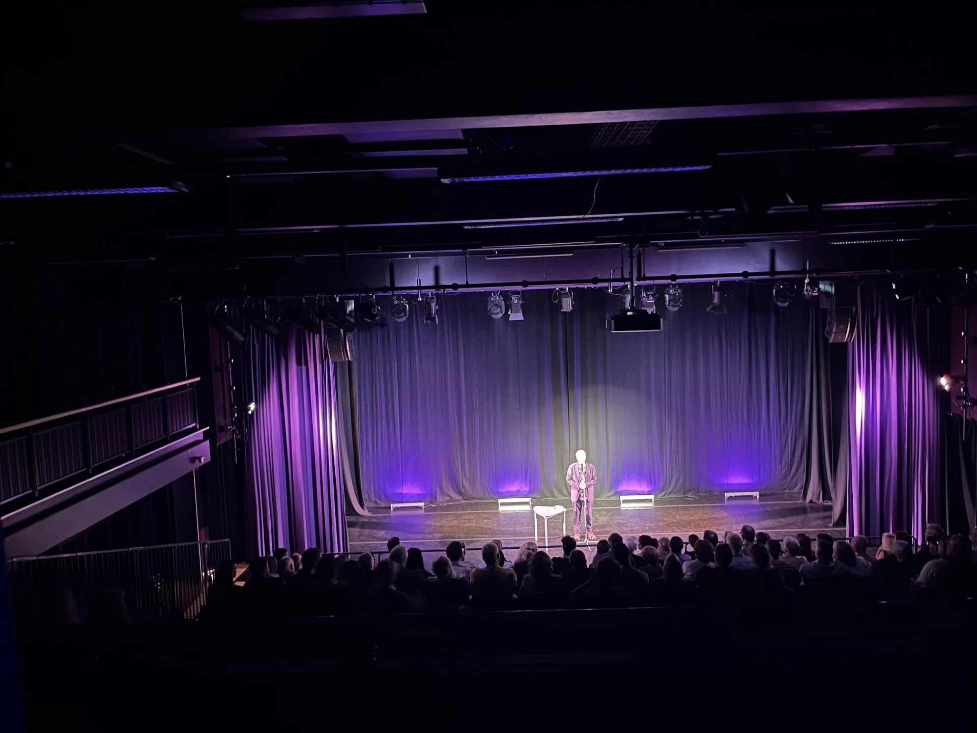 A stage photo from the top of the audience. Nigel Planer stands onstage in a spot light, purple lighting covers the stage
