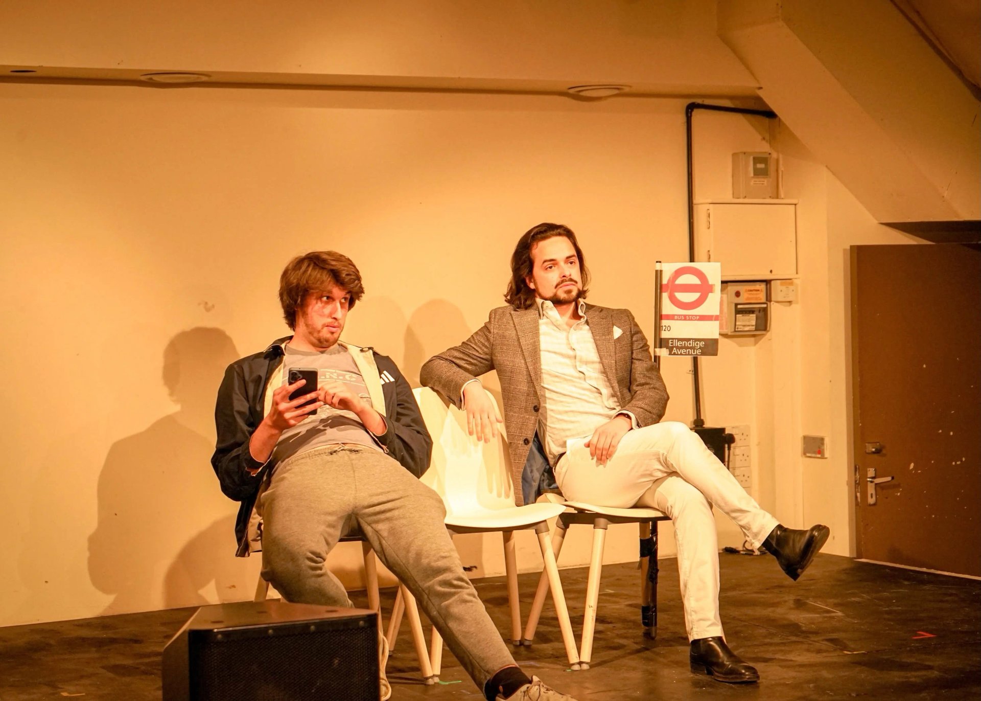 Two actors sit on chairs with a bus stop sign next to them. The one on the left looks suspicious, while the one on the right looks fed up.