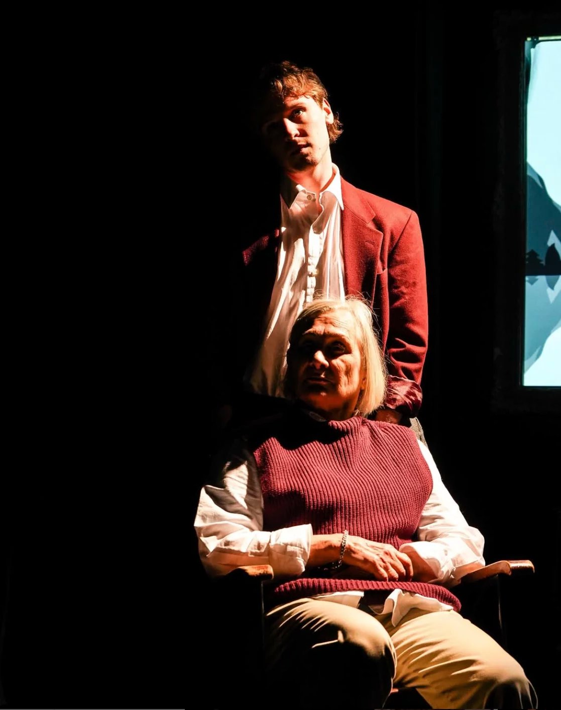 A woman sits on a chair and a man stands behind her. They are lit in a orange light that only lights the right hand side of them and leaves the rest in shadow.