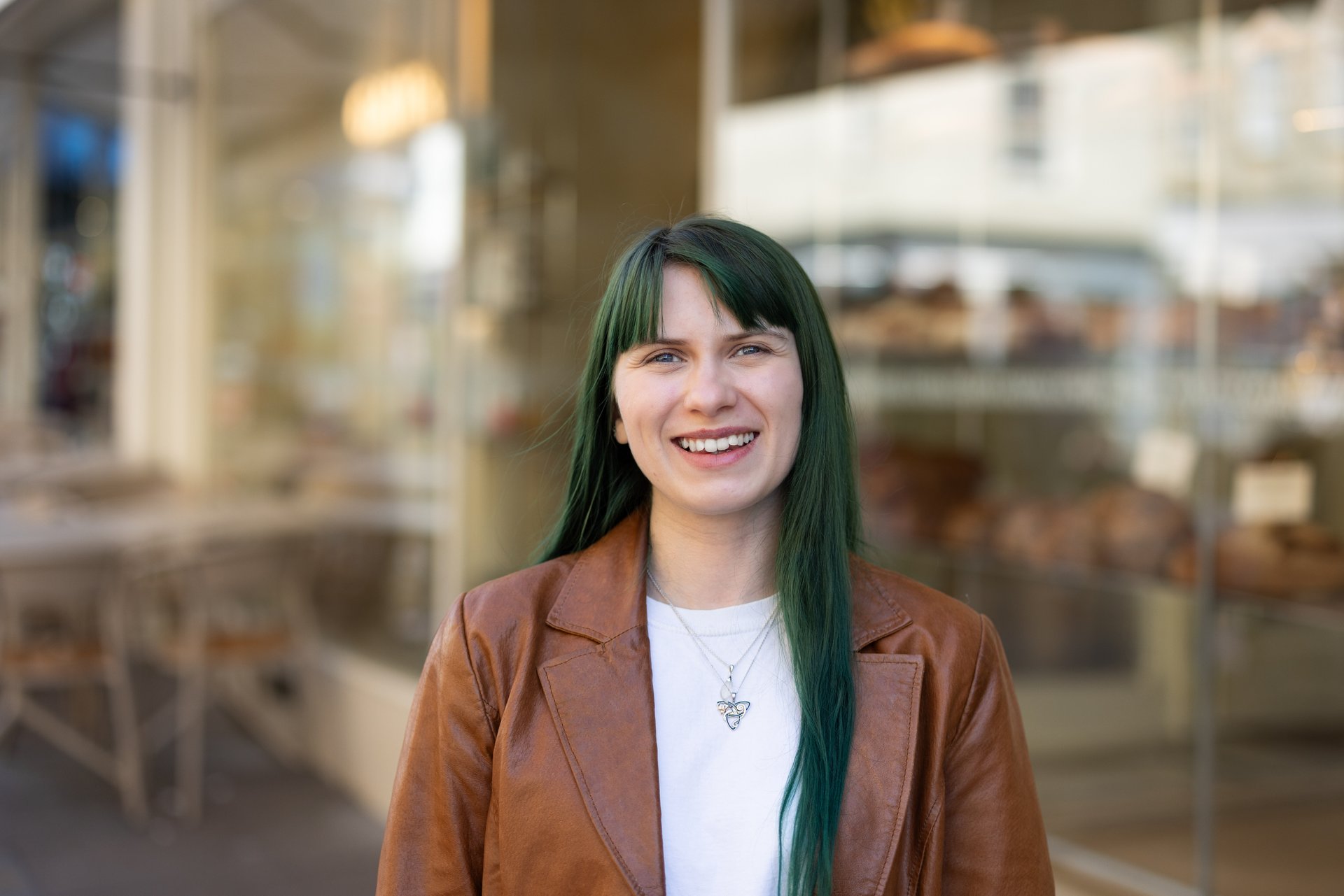 Photo of Billie-Jo Rainbird smiling. She is white with straight green hair, styled in a curtain fringe. They are wearing a white shirt and red vest.