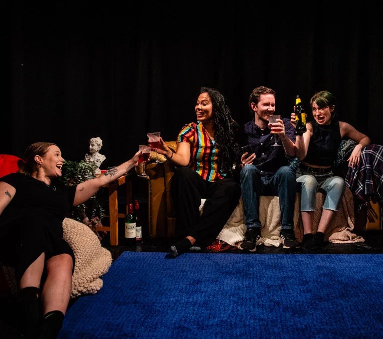Four actors on stage in a living room setting. They look happy with three sat on a sofa and one on a bean bag. They cheers each other from their seats with drinks.