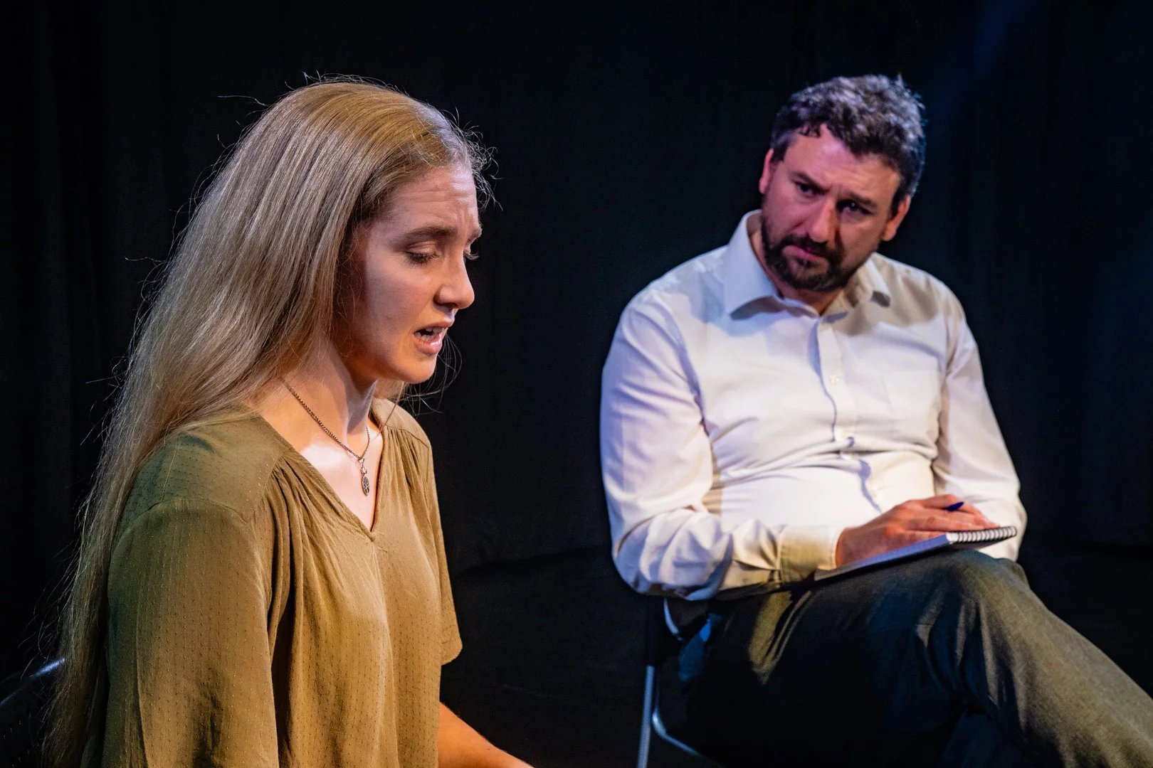 Two actors sit on chairs talking to one another. The female actor looks upset and the male looks concerned.