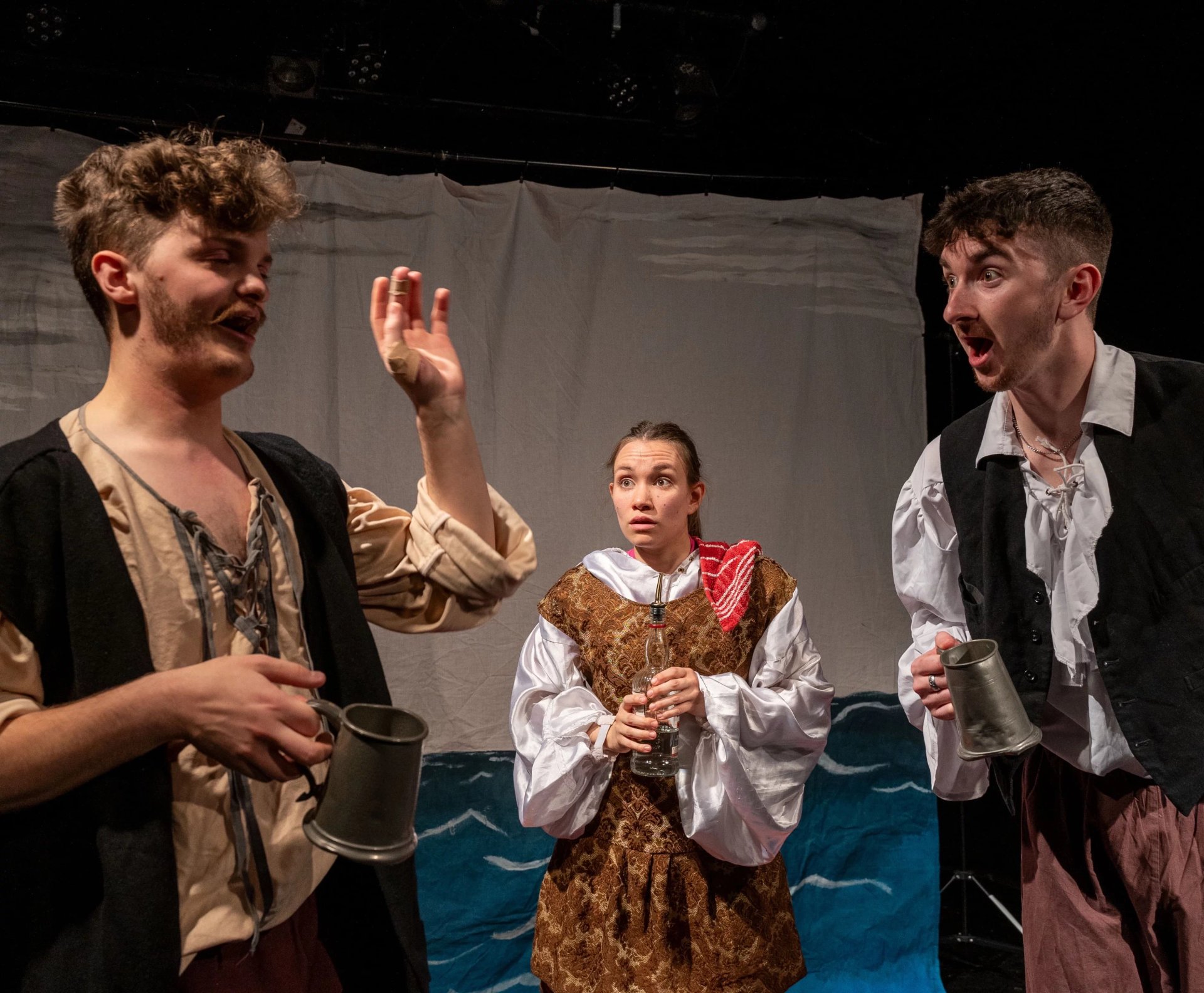 Two men look at each other in jest. A woman stands behind them looking in mild concern. The men are holding tankard. There is a sheet behind them showing the sea.