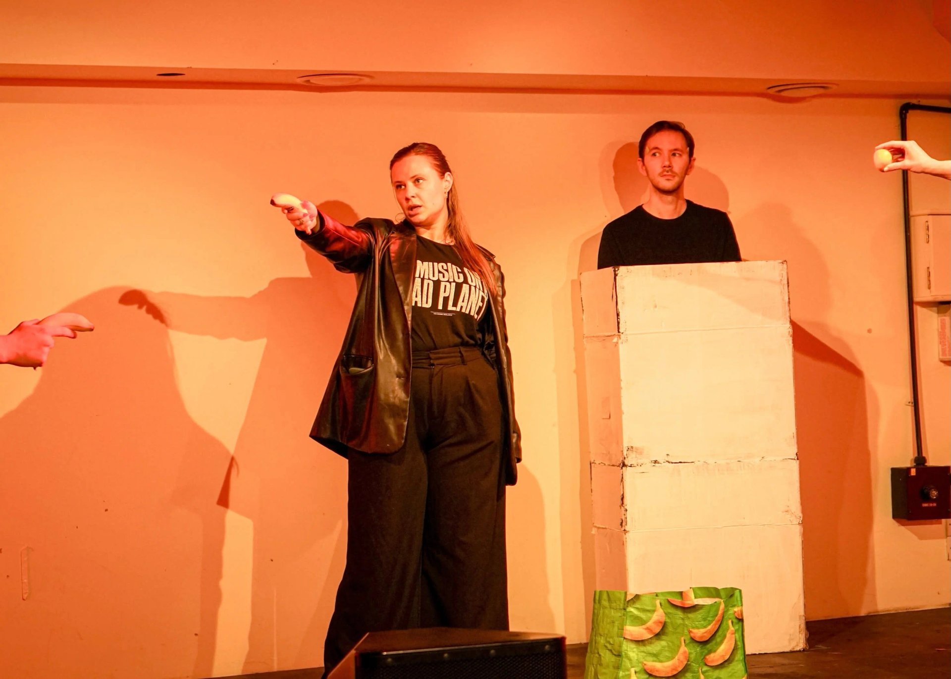 An actor in a black leather jacket points a banana like a gun. Behind them an actor stands in a white cardboard box.