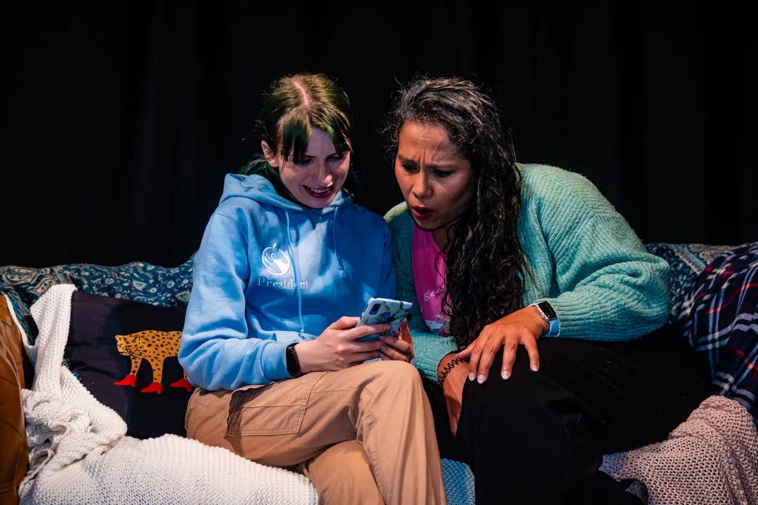 Billie-Jo as Mercy in love you, bye. She and another actor sit together looking at a phone screen with smiles on their faces.
