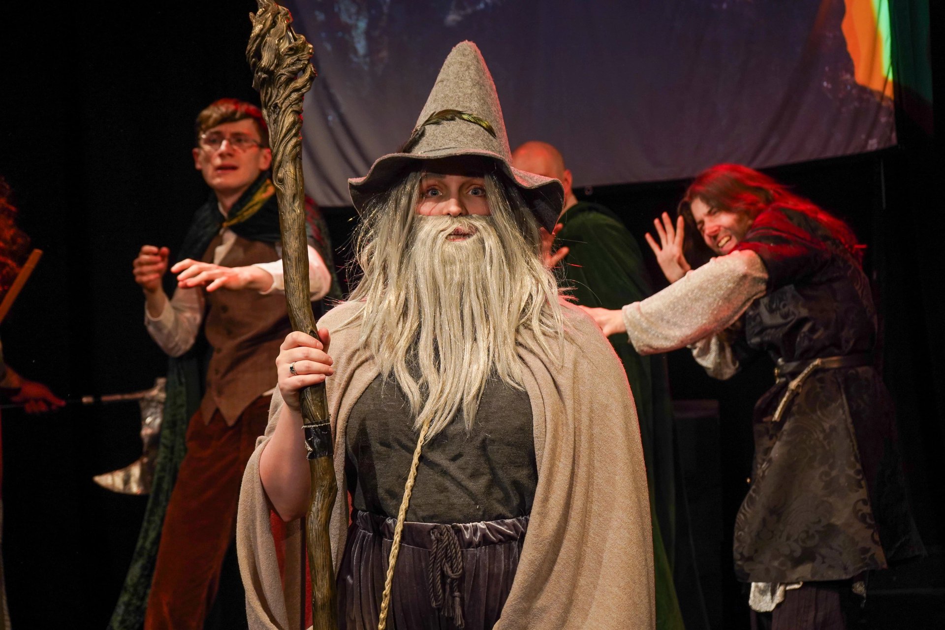 A man looks at the camera. They are wearing a grey pointed hat, grey beard and wig, grey cloak and robes. They hold a tall wooden staff. Three people stand behind them cowering in fear.