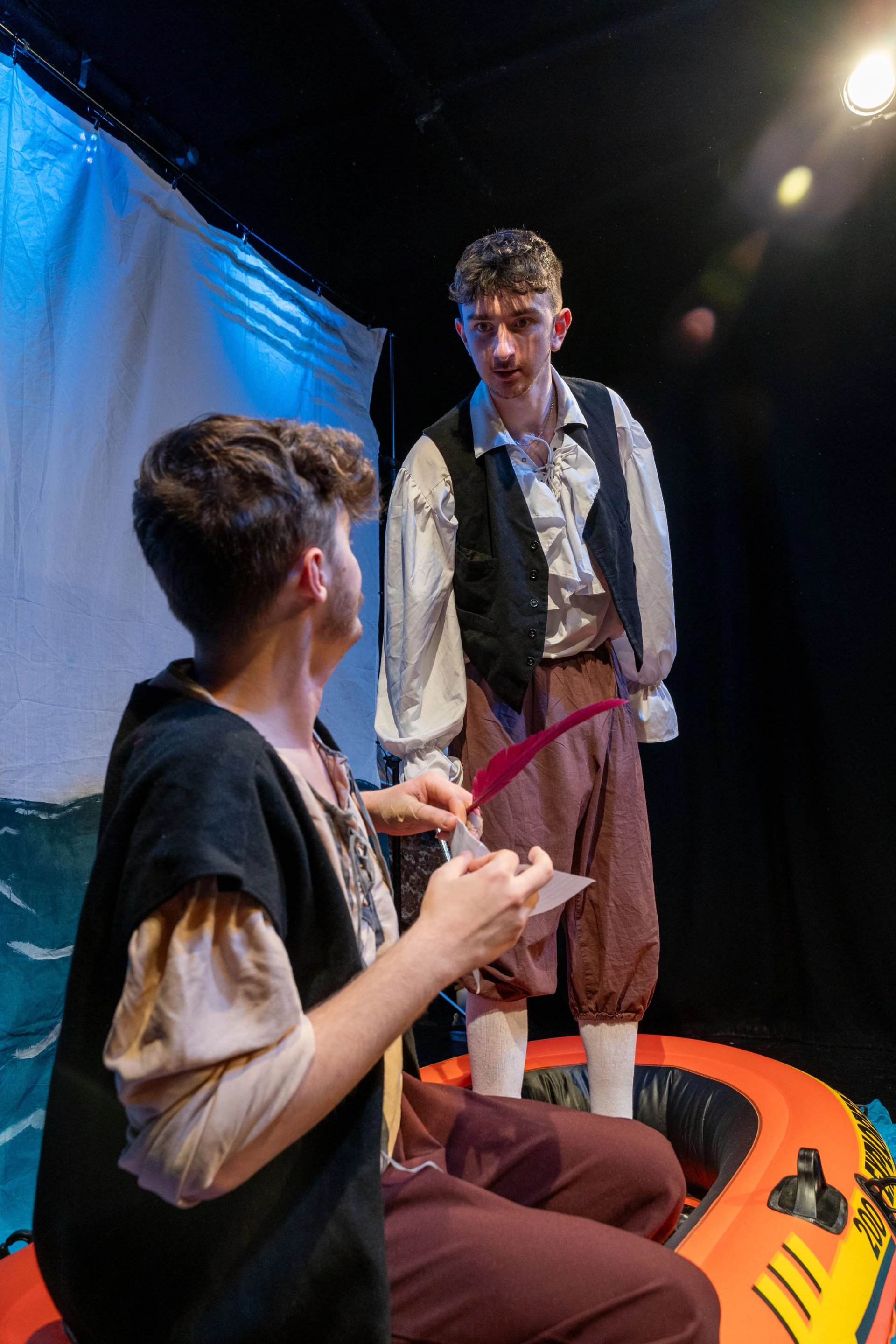 Two men in a orange life boat. One is stood up looking down at the other. They are in frilled shirts, brown trousers and black waist coats. There is a white sheet showing the seat behind them.