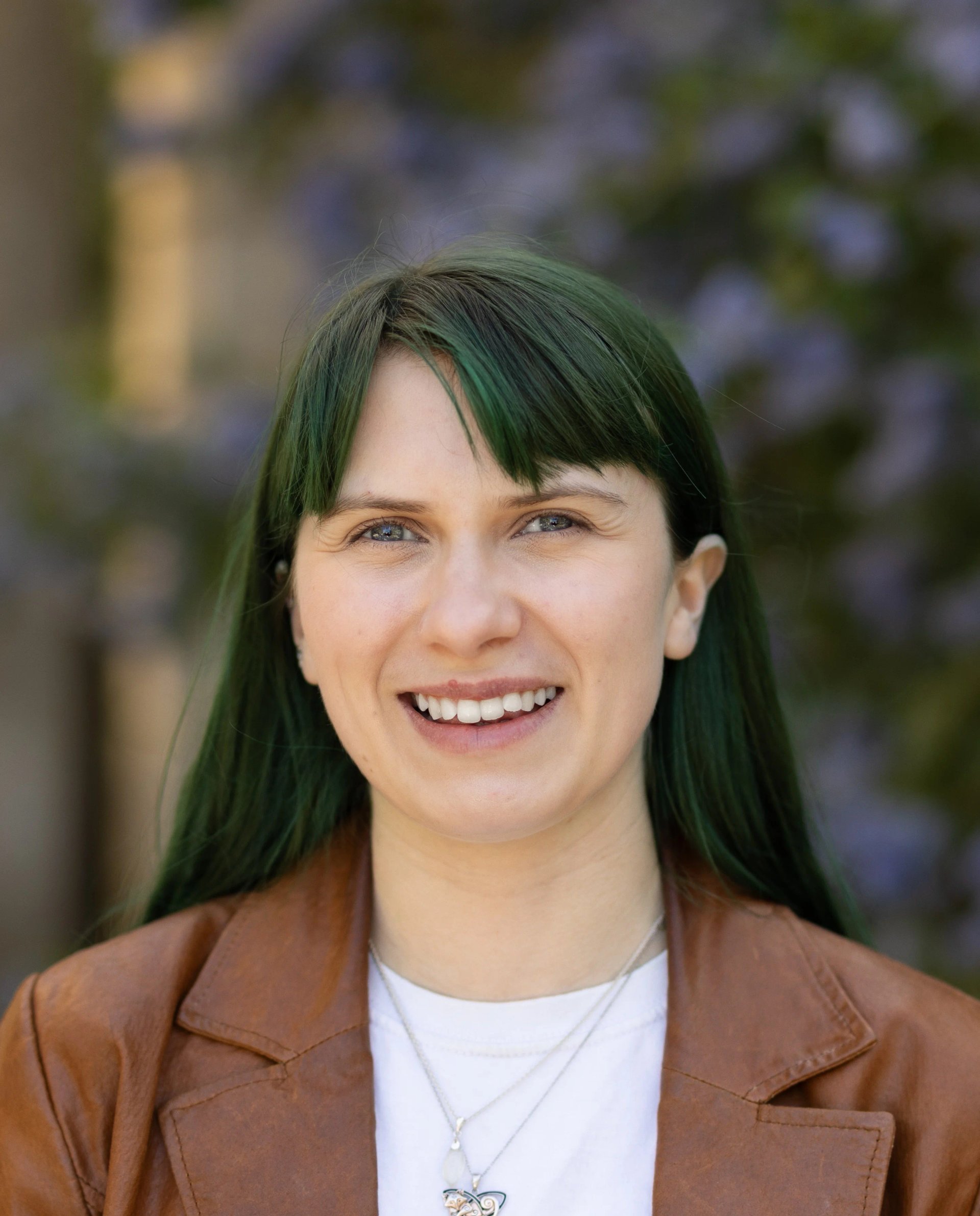 Acting headshot for Billie-Jo. Her head and shoulders are visible. She is white with blue eyes and green hair in a curtain fringe. She is wearing a white t-shirt and brown leather jacket. Behind her is an out of focused bush with purple flowers.