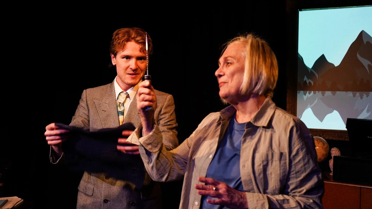 A man and woman stand looking at the knife the woman is holding. They look in awe. The woman wears a shirt and t-shirt and the man is in a suit.