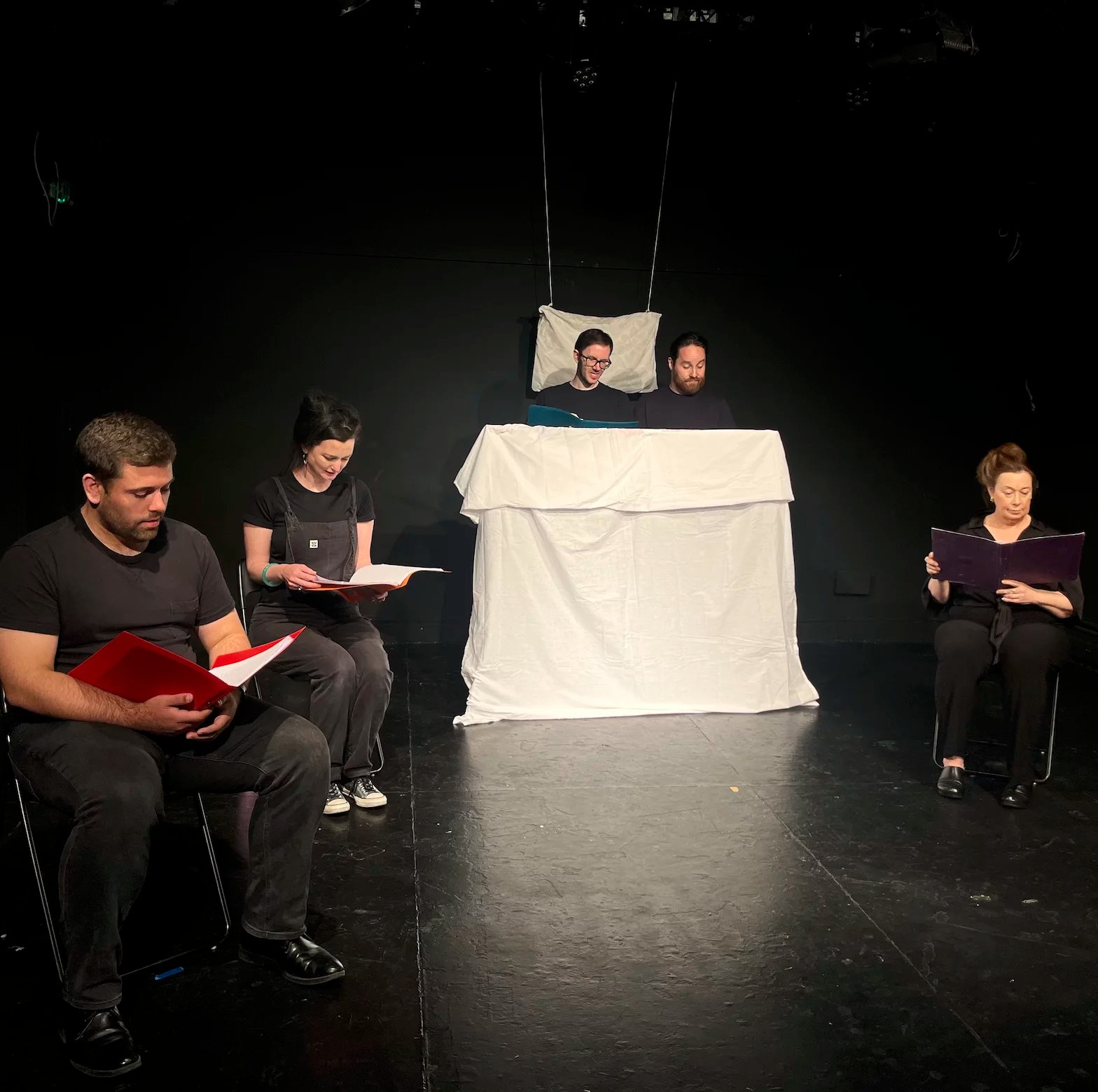 Three actors sit on chairs in a horse shoe shape. They all look at scrips in their hand. Two actors stand behind a white sheet being used as a bed