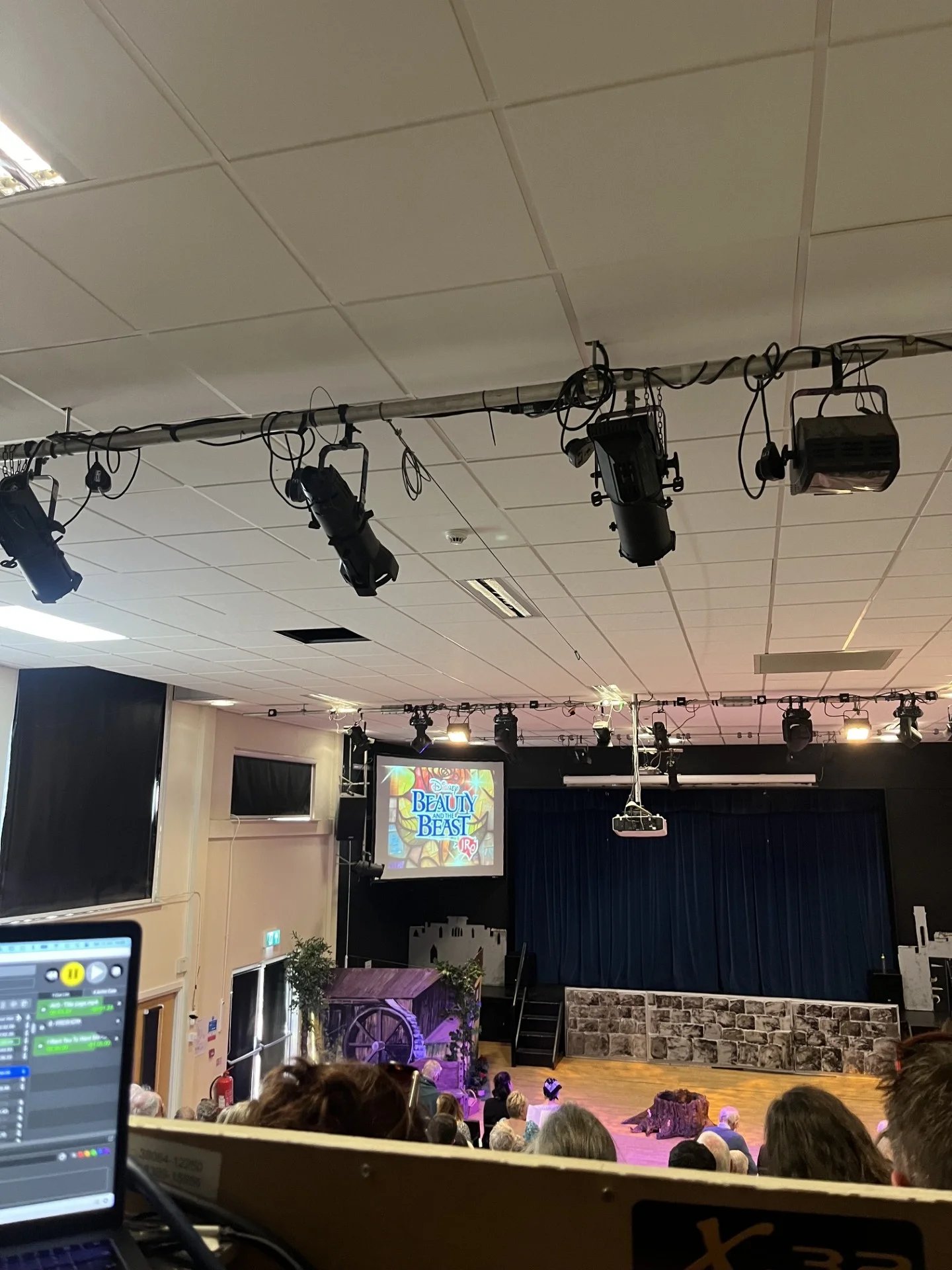 A stage photo from the back of a theatre. A projector screen says Beauty and the Beast. A cottage and stone wall are on stage covered in pink lighting.