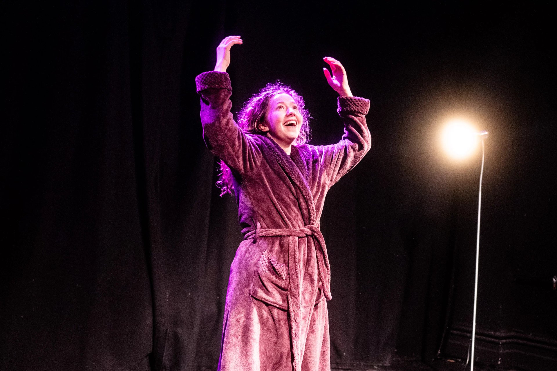 An actor in a dressing gown has their arms raised above their head. They have an elated expression and are lit in purple and yellow.