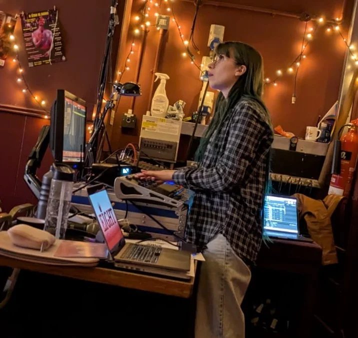 Billie-Jo Rainbird as a technician for Eight. She is standing at a tech desk working the lighting. Several laptops and computers are around her showing other systems.