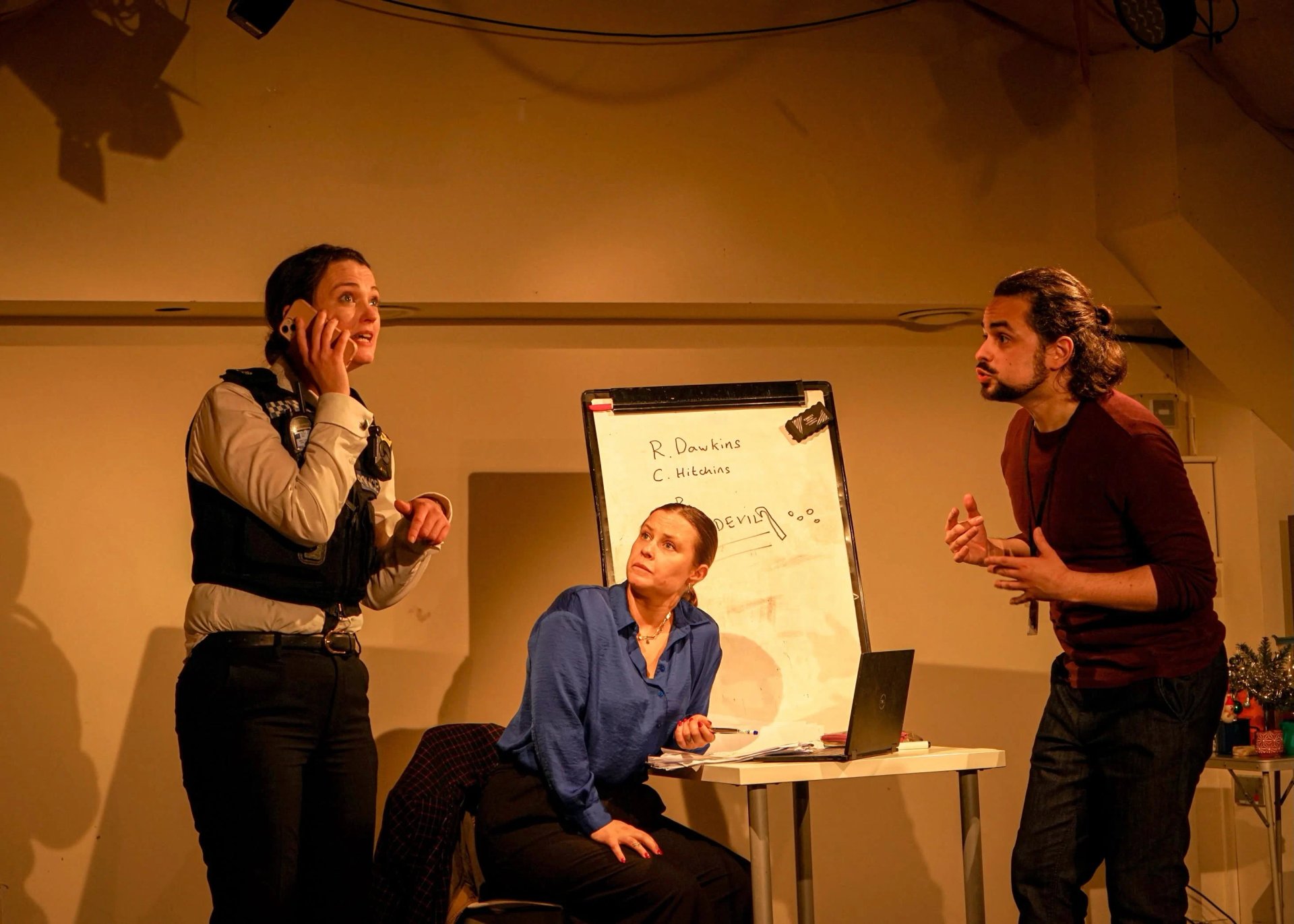An actor in a police uniform is on the phone. Behind them is another actor sat at a table looking concerned.
