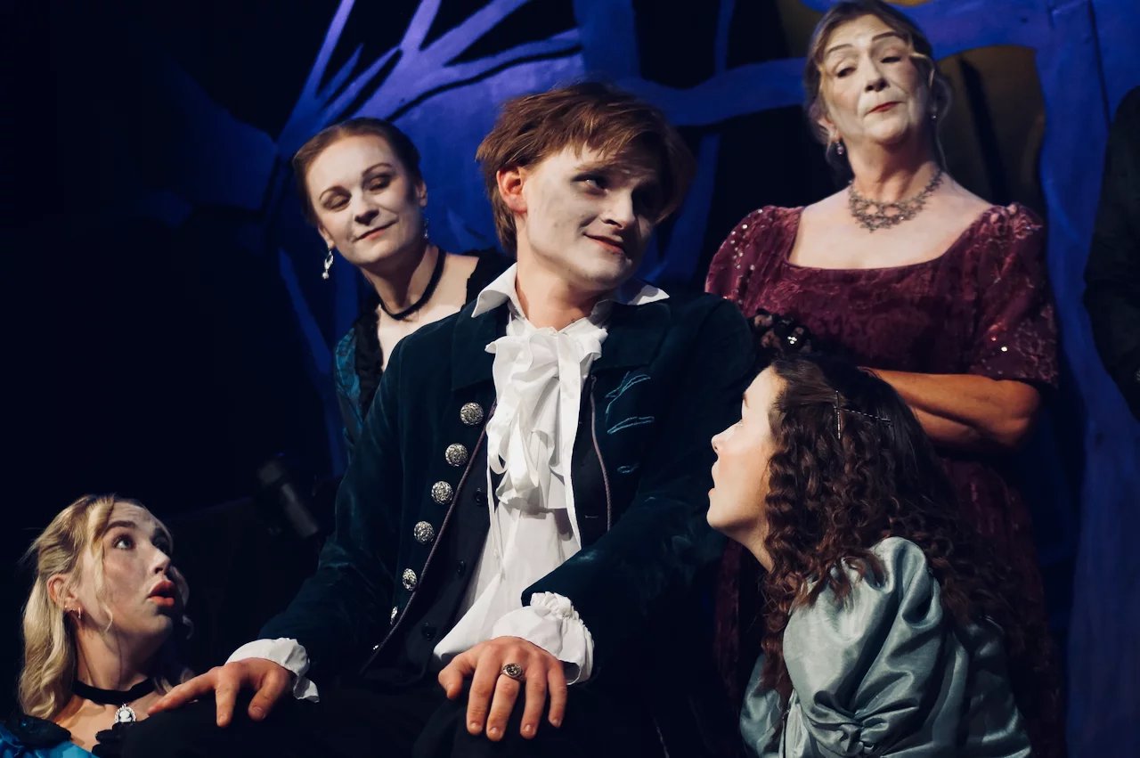 A male actor sits surrounded by female actors. They all have painted white faces and are in 1800's clothing. Behind them are tree branches lit in blue.